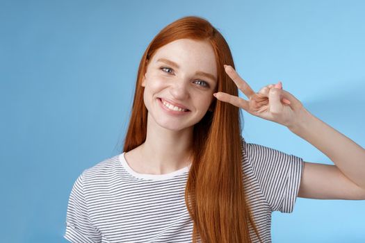 Cheerful friendly gorgeous redhead girl glancing happily show peace victory sign tilting head cute smiling broadly white teeth having fun express positivity optimism, standing blue background.