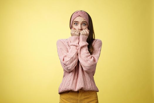 Stunned scared timid insecure cute young girl frightened gasping from fear pressing hands to mouth standing in stupor shocked, horrified stupified against yellow background being victim of abuse.
