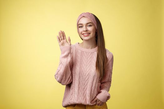 Hello adventures. Charming glamour 20s girl in sweater, knitted headband waving palm saying hi friendly smiling, holding hand in pocket gretting friends, posing positive and carefree over yellow wall.