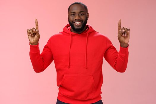 Devious cute african-american bearded guy in red hoodie smiling mysterious know exactly what you want pointing raised index fingers up grinning show perfect copy space promo, pink background.