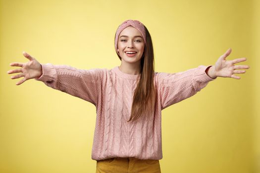 Attractive friendly positive young european woman wearing sweater, headband welcoming extanding arms and smiling at camera giving hugs, embraces friends, cuddling against yellow background. Copy space