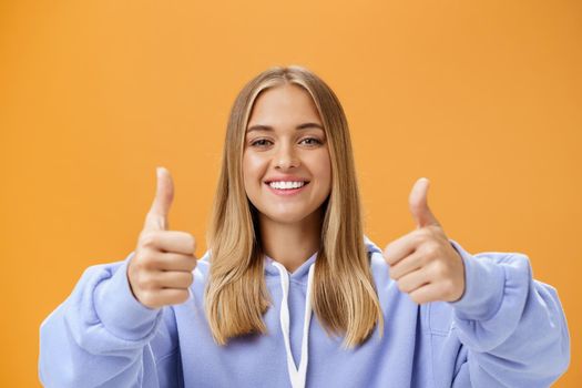 Portrait of happy optimistic woman showing thumbs up gesture and smiling supportive with cheerful look approving and liking great product giving positive opinion and recommendation over orange wall. Copy space