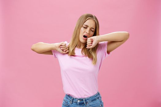 Too tired to work today. Lazy and exhausted attractive young female student doing homework all night yawning with closed eyes while stretching hands from after tiresome project over pink wall. Body language concept