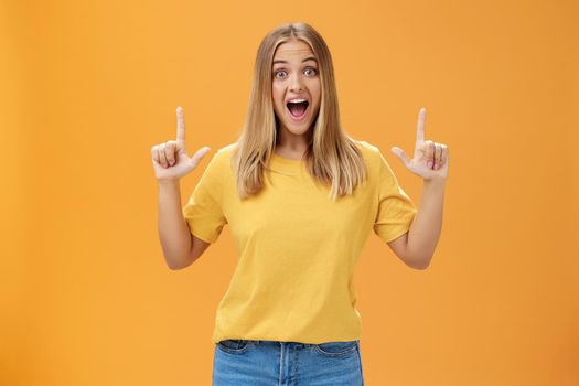 Portrait of joyful surprised and impressed delighted young woman with tanned skin and fair hair opening mouth from amazement and joy raising hands pointing up at awesome copy space against orange wall. Lifestyle.