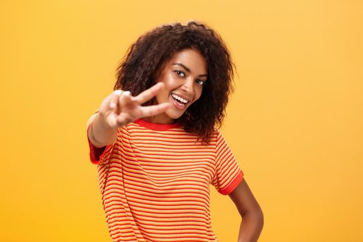 Hey peace my friend. Charming outgoing and confident carefree dark-skinned girl with afro hairstyle in trendy t-shirt pulling hand with victory gesture towards camera smiling over orange wall.