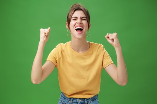 Waist-up shot of charismatic energized and excited female in yellow t-shirt closing eyes yelling from joy and happiness raising hands in cheer celebrating successful news over green background. Lifestyle.