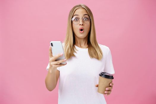 Girl receiving awesome message on smartphone while drinking coffee from cup in cafe sharing news with friend folding lips in wow sound staring surprised at camera holding cellphone over pink wall. Lifestyle.