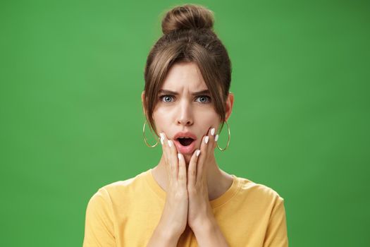 Shocked woman reacting to pimple looking disturbed and displeased in mirror holding hands pressed to cheeks open mouth and frowning being disappointed and upset over green background. Body language and emotions concept