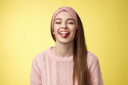 Positive entertained cute funny glamour young european girl in sweater, knitted trendy headband smiling fooling around showing tongue playfully, mocking friend enjoying sunny day over yellow wall.
