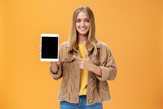 Get ready for autumn semister with new digital tablet. Portrait of charming female in corduroy jacket showing gadget screen pointing at device and smiling pleased and friendly over orange wall. Technology and advertising concept