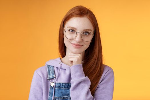 Confident happy relaxed european redhead skillful young female entrepreneur glasses establish startup smiling delighted look pleased result touch chin thoughtful smart, orange background.