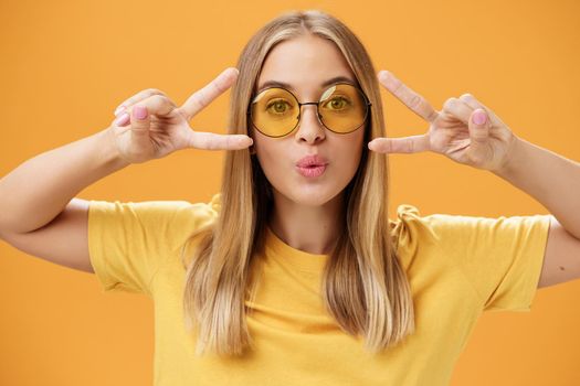 Carefree charismatic pretty woman in trendy round sunglasses and t-shirt folding lips in kiss showing peace or disco signs near eyes dancing having fun on party against orange background. Lifestyle.