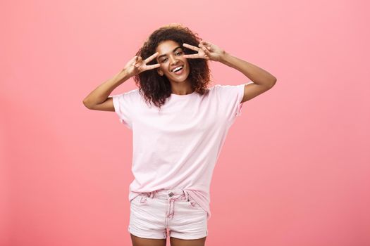 Indoor shot of charismatic playful dark-skinned woman with afro hairstyle tilting head showing peace or victory signs over eyes and smiling feeling amused chilling on awesome party over pink wall. Lifestyle.