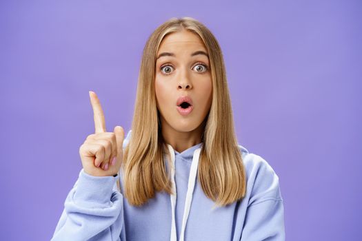 Oh I know idea. Excited cute young woman with tan in hoodie raising index finger in eureka gesture folding opened lips and staring amazed at camera having suggestion what do over blue background. Copy space