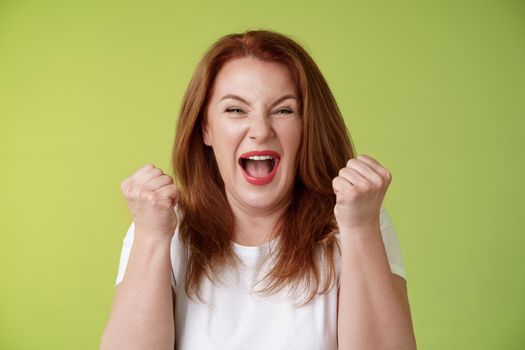 Yeah we did it. Joyful lucky redhead middle-aged female winner pump fists up celebration success gesture yelling triumph joy smiling broadly celebrate awesome news stand green background happy.