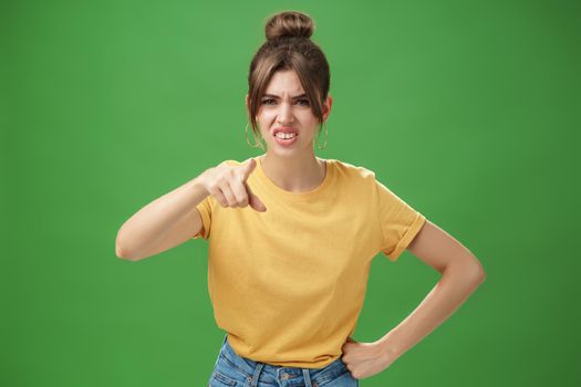 Portrait of arrogant impolite young woman in yellow t-shirt pointing at camera squinting and grimacing from disdain and scorn standing unimpressed judging against green background. Copy space
