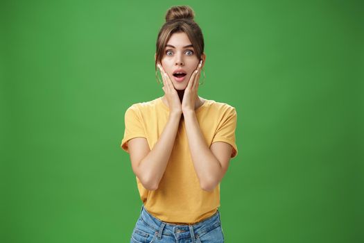 Woman feeling surprised and delighted enjoying good result after detox and applying facial skincare mask touching cheeks opening mouth from amazement and excited posing over green background. Lifestyle.