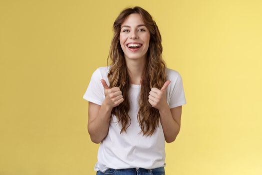Pleased outgoing cheerful good-looking caucasian girl curly long haircut white t-shirt show thumbs up smiling lively pleased like awesome performance approve good choice yellow background. Lifestyle.