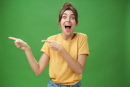 Waist-up shot of enthusiastic and charismatic impressed happy girl laughing and smiling broadly pointing left at funny hilarious thing reacting surprised and entertained, posing over green background.