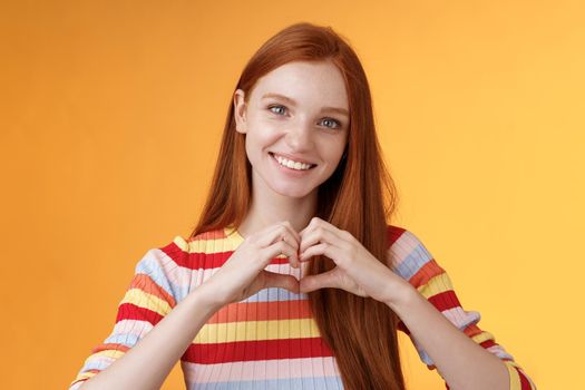 Lovely friendly-looking charismatic smiling redhead girl express affection love and friendship grinning show heart sign chest showing sympathy, confess romantic feelings, standing orange background.