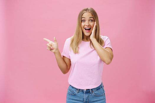 Portrait of surprised enthusiastic and charismatic tanned cute woman with fair straight hair touching cheek from joy and amazement pointing left delighted and amused over pink background. Copy space