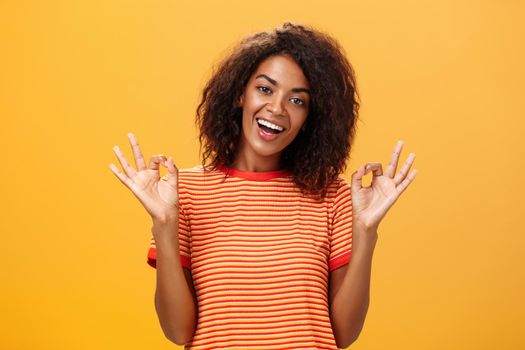 Waist-up shot of chill outgoing calm girlfriend with confident look in trendy striped t-shirt showing okay or excellent gestur and smiling broadly assuring everything alright over orange background. Lifestyle.
