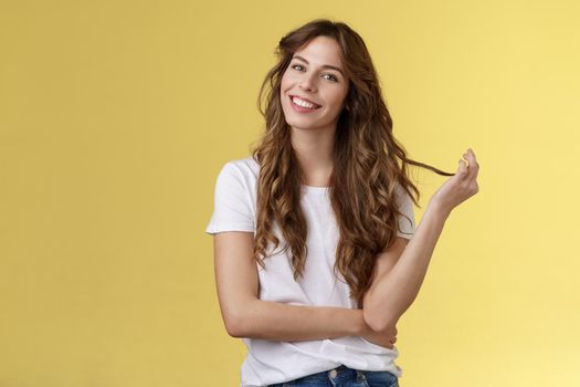 Silly cute feminine sensual curly-haired woman tilt head playing hair strand rolling curl smiling delighted interested listening flirty conversation coquettish gazing camera stand yellow background. Lifestyle.