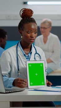 Medic showing green screen on tablet to senior man sitting at desk in healthcare office at hospital. Doctor vertically holding isolated template with mockup background and chroma key.