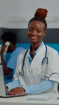 Close up of woman with doctor occupation using laptop technology on desk at clinic. Portrait of medic looking at camera and typing on device keyboard while having white coat and stethoscope