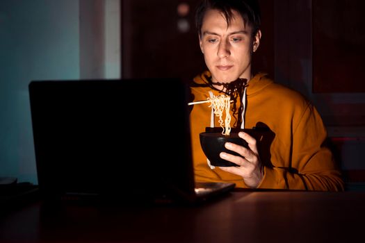 A young man in a yellow casual hoodie eats noodles with sticks and watches a movie at night on a laptop at home, a man works, studies and has a snack.