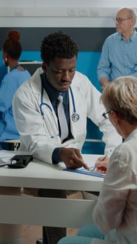 Adult working as doctor in white coat talking to old patient at desk in medical cabinet. Sick elder woman using pen for signature on examination files sitting with young specialist