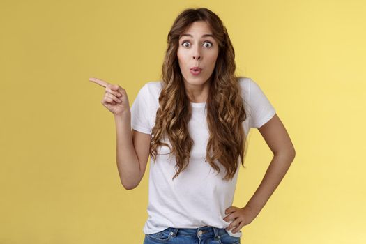 Wow incredible lets click. Impressed surprised pretty european woman long curly hairstyle folding lips intrigued stare astonished wondered awesome cool promo offer pointing left yellow background.