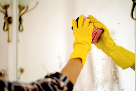 A young girl is cleaning the bathroom, applying detergent with a spray and washing the mirror with a sponge in yellow gloves on her hands. Smiling woman taking care of the cleanliness of her home.