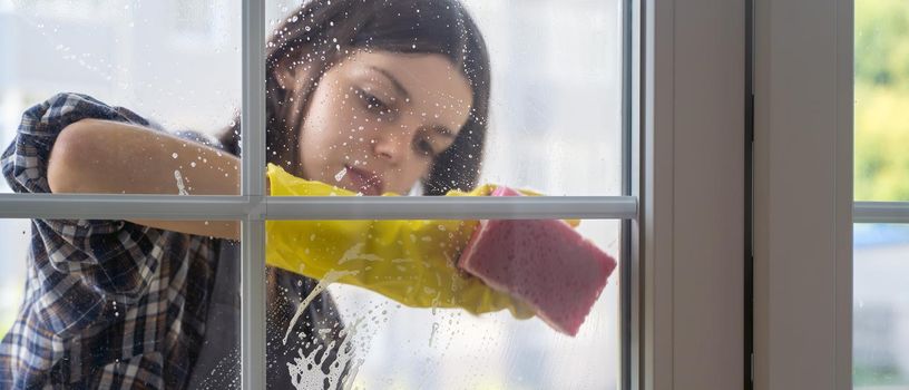 A young girl in an apron does housework, washes the windows with a detergent, wipes it dry with a rag, a woman washes the glass with a sponge.