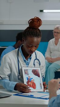Woman cardiologist holding modern tablet with cardiovascular system, explaining heart disease to elder caucasian patient looking at device, sitting in hospital room at facility