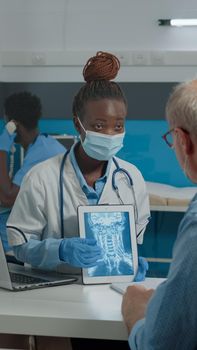 Young doctor analyzing x ray on digital tablet for old patient with disease sitting at desk with plexiglass wall. Medic wearing face mask, explaining radiography to senior man
