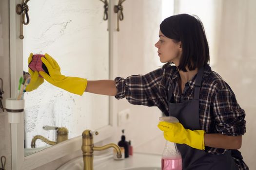 A young girl is cleaning the bathroom, applying detergent with a spray and washing the mirror with a sponge in yellow gloves on her hands. Smiling woman taking care of the cleanliness of her home.