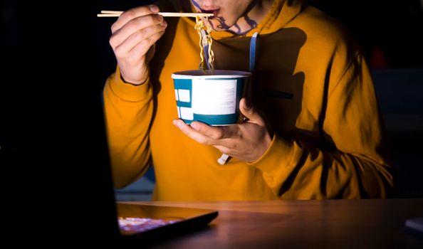 A young man in a yellow casual hoodie eats noodles with sticks and watches a movie at night on a laptop at home, a man works, studies and has a snack.