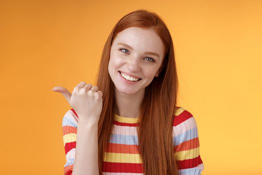 Friendly tender feminine kind redhead woman blue eyes pointing thumb left showing location where find product copy space gladly help discuss interesting promo grinning flirty, orange background.