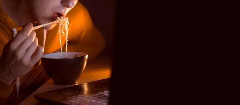 A young man in a yellow casual hoodie eats noodles with sticks and watches a movie at night on a laptop at home, a man works, studies and has a snack, close up view and dark copy space.