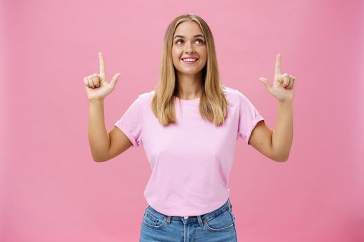Girl daydreaming pointing and looking with upbeat and admiring expression up smiling cheerfully enjoying nice sunny weather walking in casual t-shirt and jeans posing against pink background. Copy space