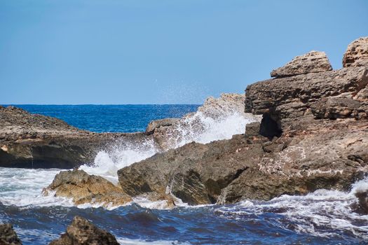 Rocks on the shore hit by the waves. Balearic Islands, Mediterranean Sea, blue sky, splashes of water.