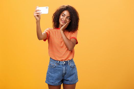 Woman made make-up all day to post new photo online taking selfie on brand new smartphone making cute face leaning head on palm smiling gently at device screen posing over orange background. Technology and lifestyle concept