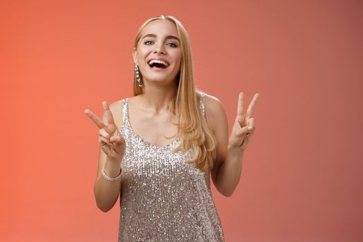 Funny carefree charming caucasian blond female model in silver stylish glamour dress show peace victory gesture smiling broadly have fun celebrating birthday party, standing red background.