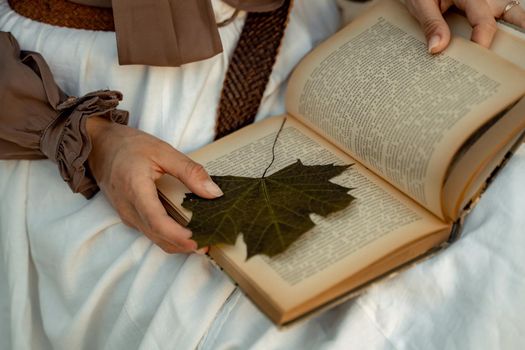 A woman in a white skirt and brown blouse sits and holds an open book in her hands. She is reading a book. There is a maple leaf in the knit as a bookmark