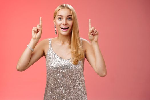 Charming amazed blond european woman in fabulous silver glittering dress raise hands point up amused enjoying watching shooting stars, fireworks gaze camera excited happy surprised, red background.