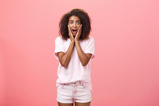 Worried, shocked african american female friend with curly hairstyle in trendy outfit being on vacation seeing scary animal shouting and gasping with opened mouth holding hands on face over pink wall.