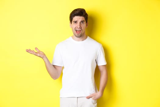 Confused and shocked man complaining, raising one hand and looking bothered, standing near yellow copy space.