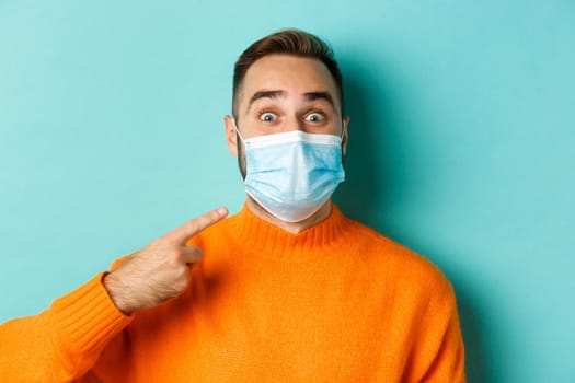 Covid-19, social distancing and quarantine concept. Close-up of cheerful man pointing finger at his medical mask, standing over turquoise background.