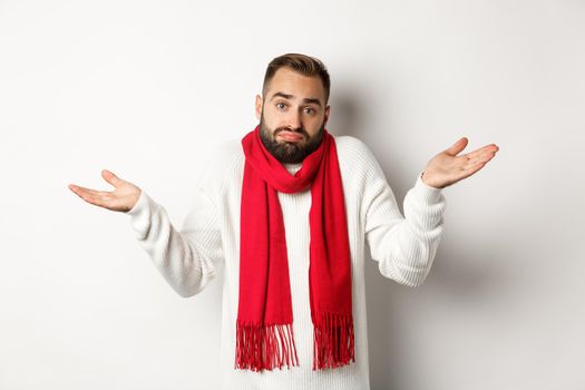 Confused bearded man shrugging, raising hands up and looking clueless, dont know anything, standing in sweater and christmas scarf, white background.